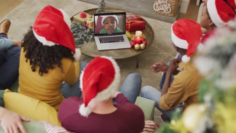 Diverse-family-with-santa-hats-using-laptop-for-christmas-video-call-with-smiling-boy-on-screen