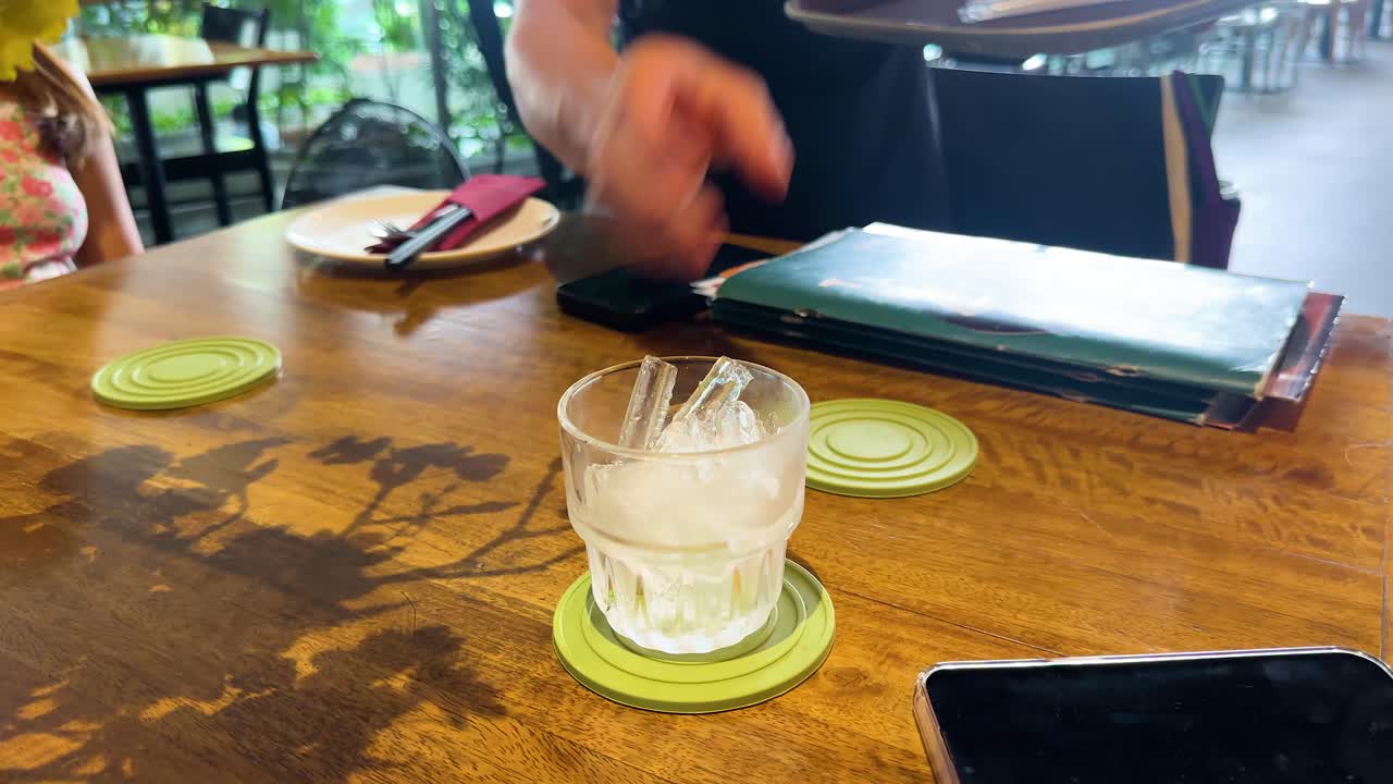 A hand places a glass of ice on a wooden table in a Bangkok restaurant, captured in natural lighting