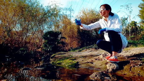 a young woman scientist at a creek, wearing protective eyewear and a lab coat, taking a water sample