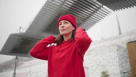 mujer de negocios con capucha roja ajusta su gorra mientras sonríe y mira a la distancia, el fondo incluye dosel moderno, colina nevada y postes de luz en un clima de invierno nebuloso