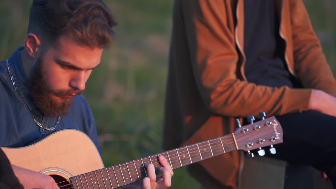 Young bearded man playing guitar while sitting on the ground. Male musician surrounded by friends at the blurred nature backdrop.