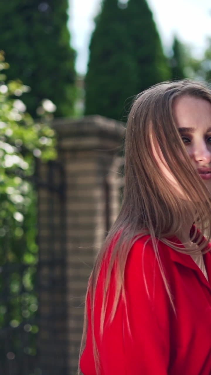 Passionate young girl posing to camera. Beautiful young model in red dress with long hair on the green trees background. Sexy woman turning around herself joyfully. Vertical video