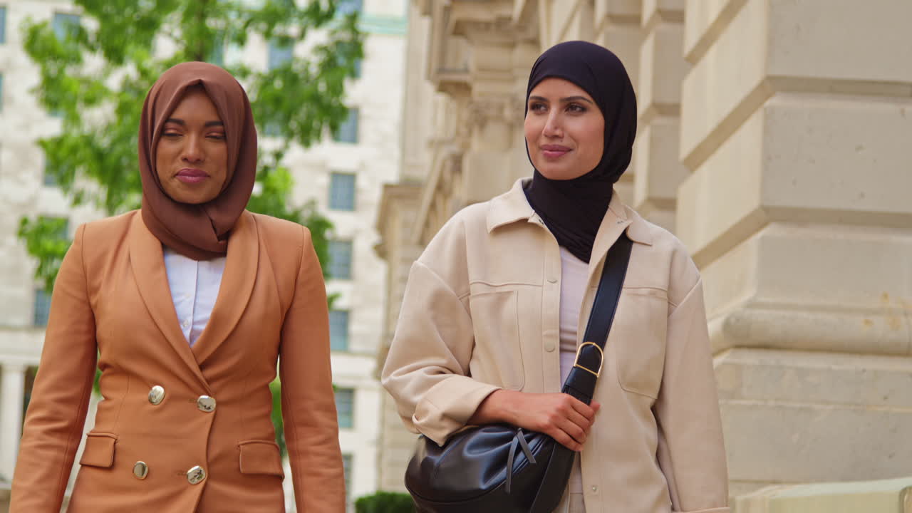 현대적인 비즈니스 슈트를 입은 히자브를 입은 두 명의 무슬림 사업가 여성이 도시 사무실 건물 4을 지나 일하러 고 있습니다.