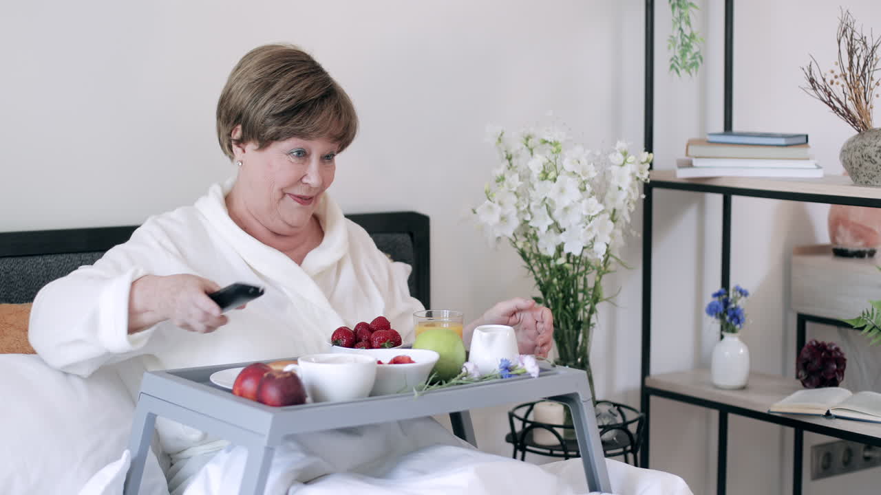 Una donna anziana felice si gusta la colazione a letto guardando la TV
