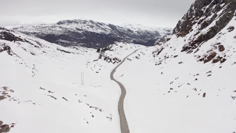 Verlassene-Straße-Zwischen-Hohen-Bergen,-Die-An-Einem-Bewölkten-Tag-In-Norwegen-Mit-Weißem-Schnee-Bedeckt-Sind