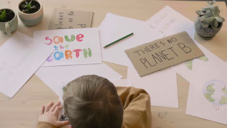 top view of blond kid drawing with green pencil sitting at a table in a craft workshop where are signs with environmental quotes