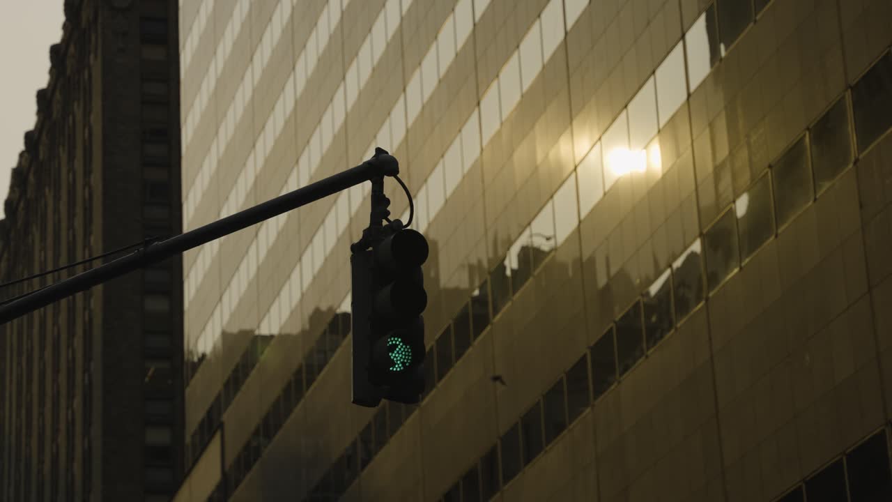 A medium shot of a traffic light with the warm glow of the sunset reflecting on the windows of a modern glass building in New York City. A contrast of urban infrastructure and evening light.