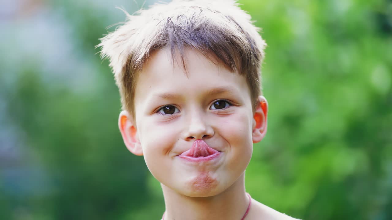 Close-up portrait of boy's face outdoors. Curious child looking at camera and showing his tongue on blur green background. Little boy tries to touch the nose by his tongue.