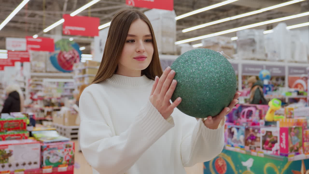 Lady holding a green shimmering Christmas ball decor, admiring it in a beautifully decorated store, with other shoppers visible in the background