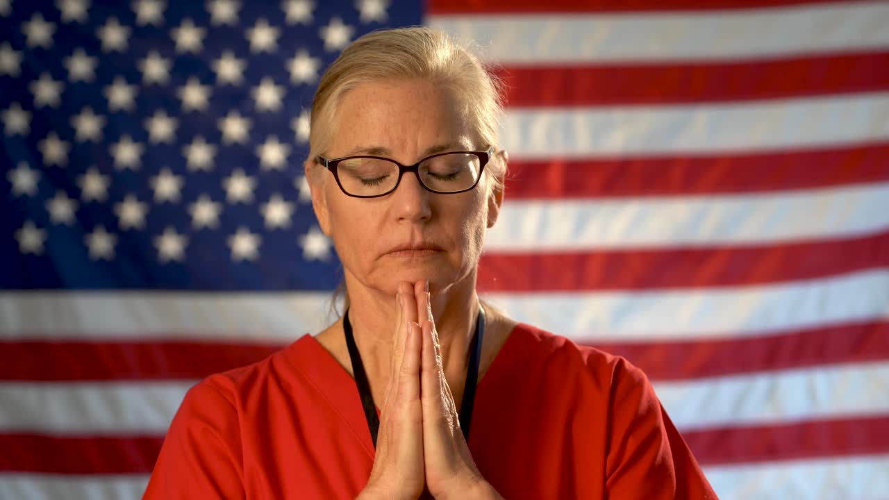 retrato medio ajustado de una enfermera de atención médica que parece preocupada y reza con una bandera estadounidense desenfocada