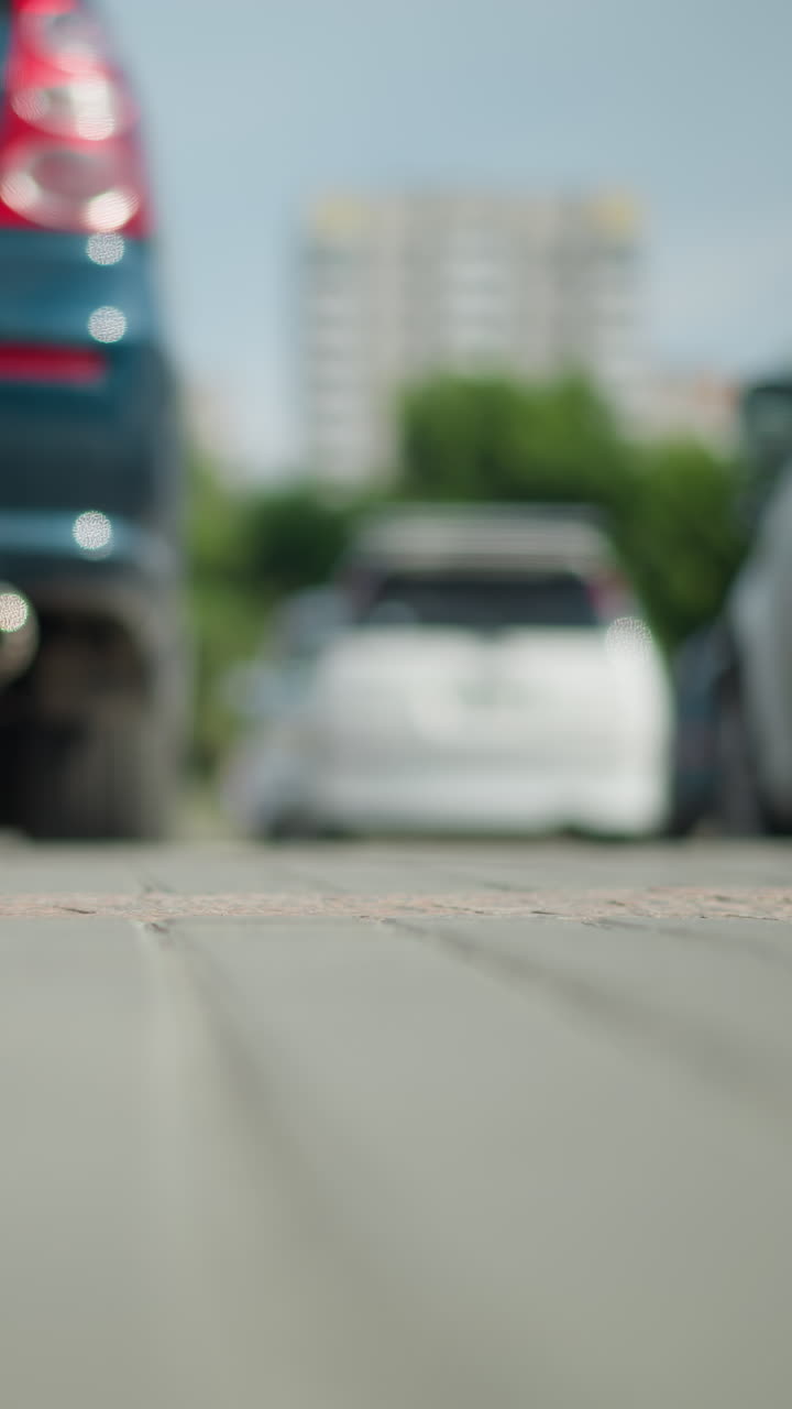 주차된 차량의 낮은 각도 시야에서 색 캔버스 스니커를 입은 사람이 지나가며, 땅에 그림자를 던지고, 먼 건물과 초록색이 흐릿한 배경에서 보입니다.