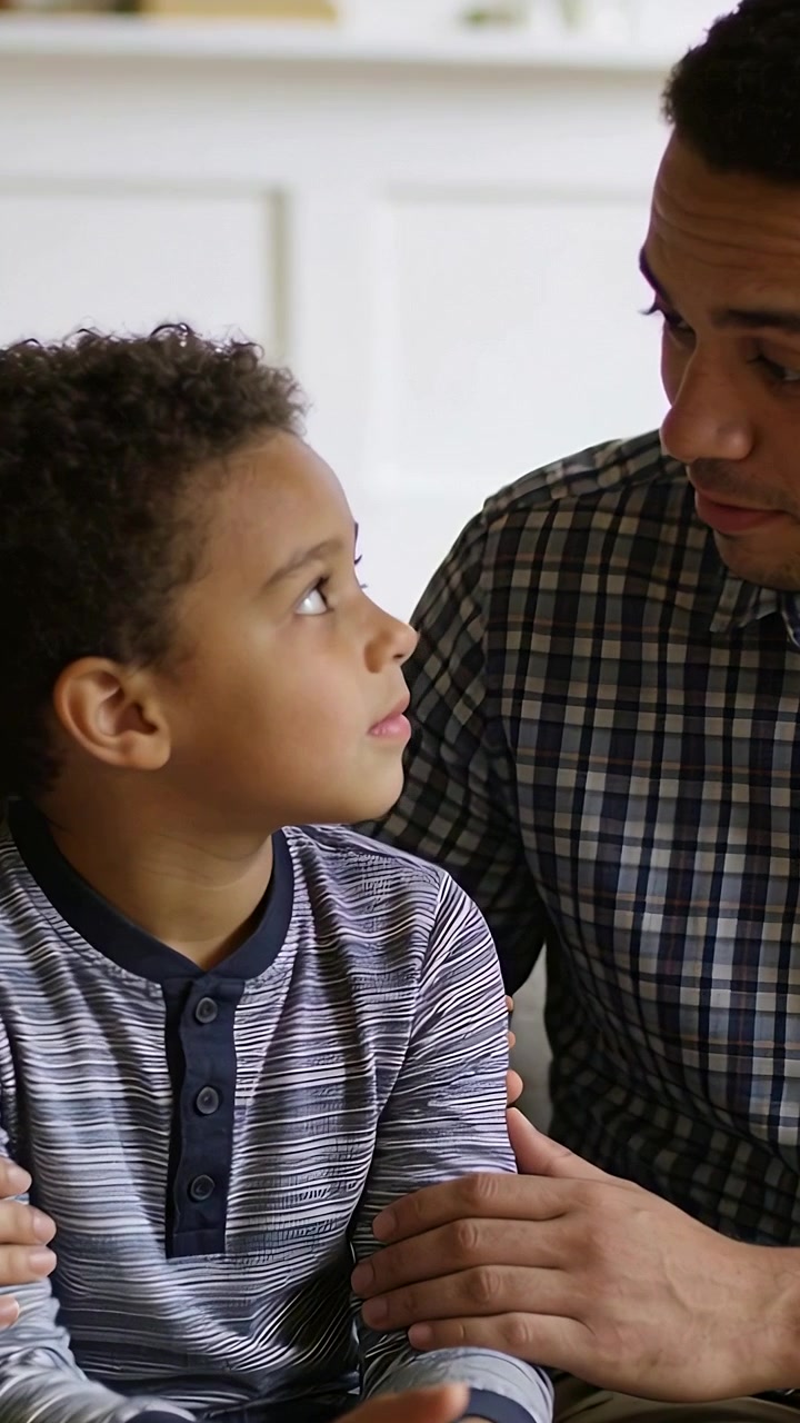 Vertical video: Son looking up father leaning touching arm on couch at home reassuring in henley