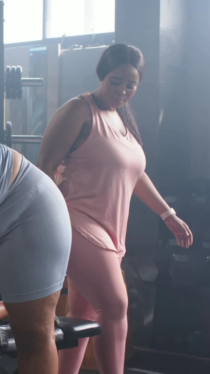 Video verticale: Donna con top rosa che dà un'indicazione e guida l'amica che fa rematore su panca in palestra per il fitness.