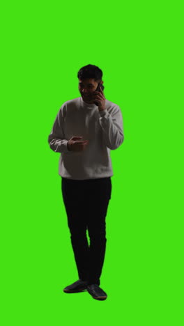 Vertical-Video-Full-Length-Shot-Of-Young-Man-Making-Call-On-Mobile-Phone-Standing-Against-Green-Screen-With-Low-Key-Lighting