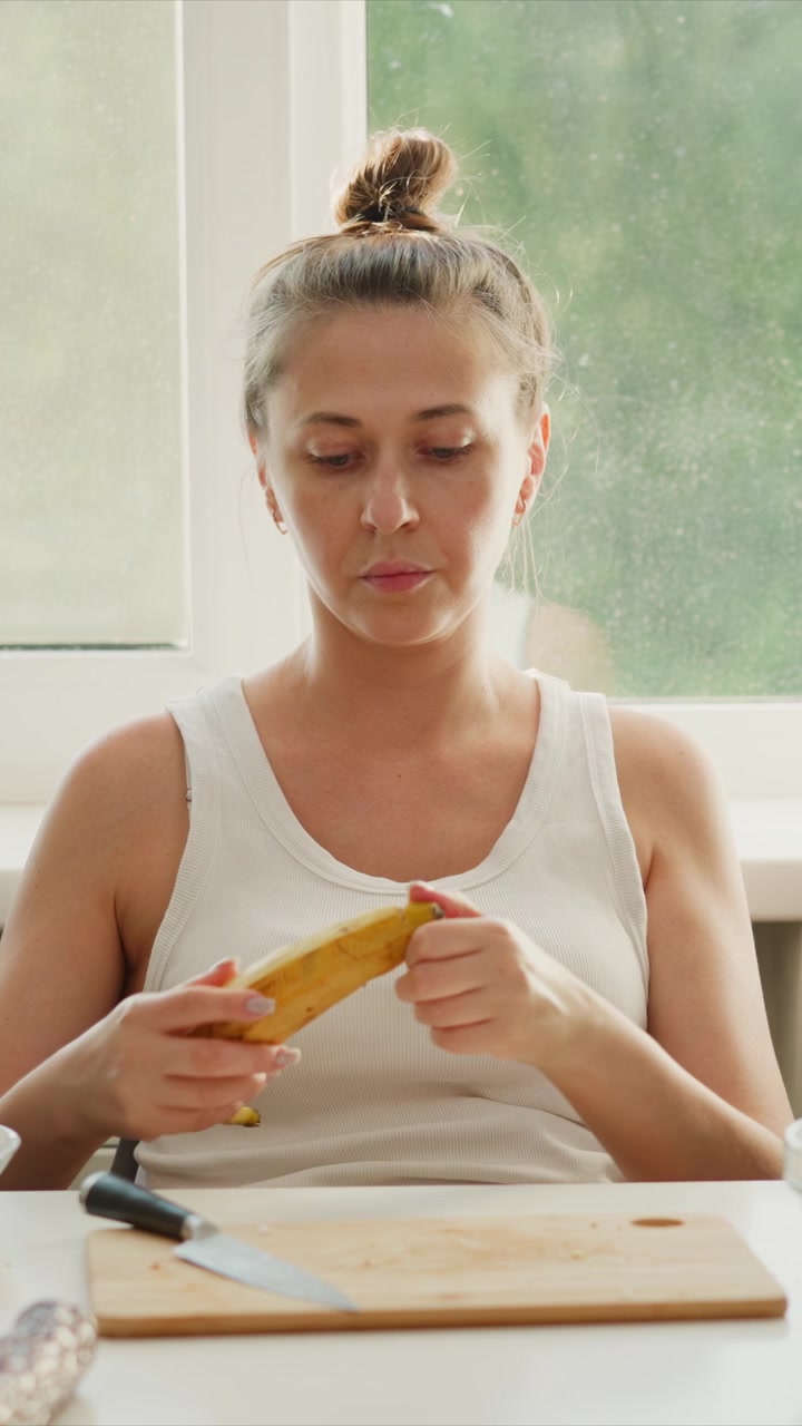Donna in canotta bianca prepara il cibo tagliando e sbucciando una banana sul bancone della cucina. Vari ingredienti come salmone, noci e ciotole sono visibili sul tavolo mentre la donna lavora sotto una luce naturale soffusa.
