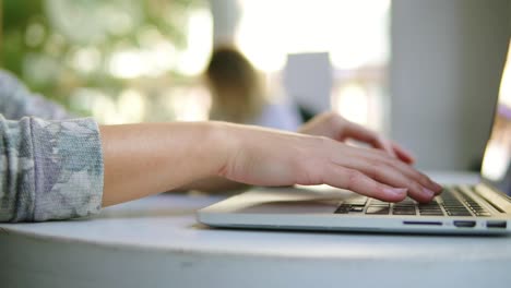 Vista-De-Cerca-De-Manos-Femeninas-Trabajando-En-Una-Computadora-Portátil.-La-Mujer-Está-Sentada-Cerca-De-La-Ventana-En-La-Cafetería-Y-Escribiendo.-Taza-Blanca-Con