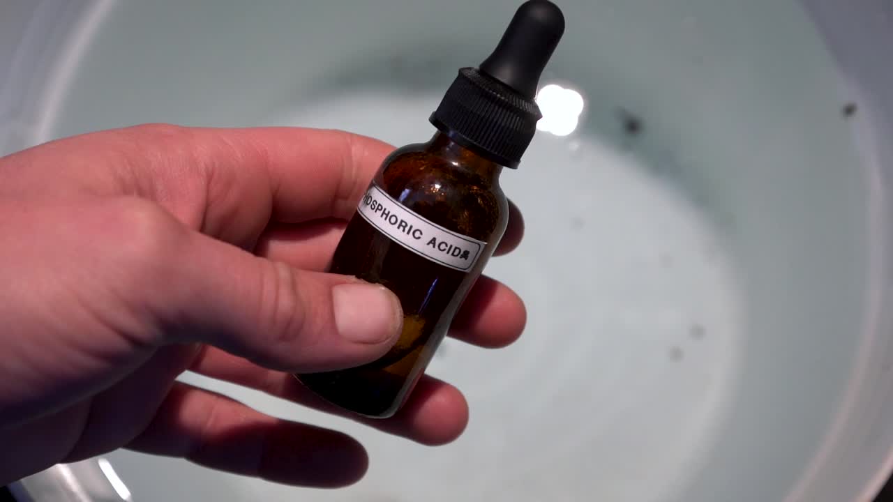 mano sujetando una pequeña botella de ácido fosfórico sobre un balde de agua - primer plano, punto de vista