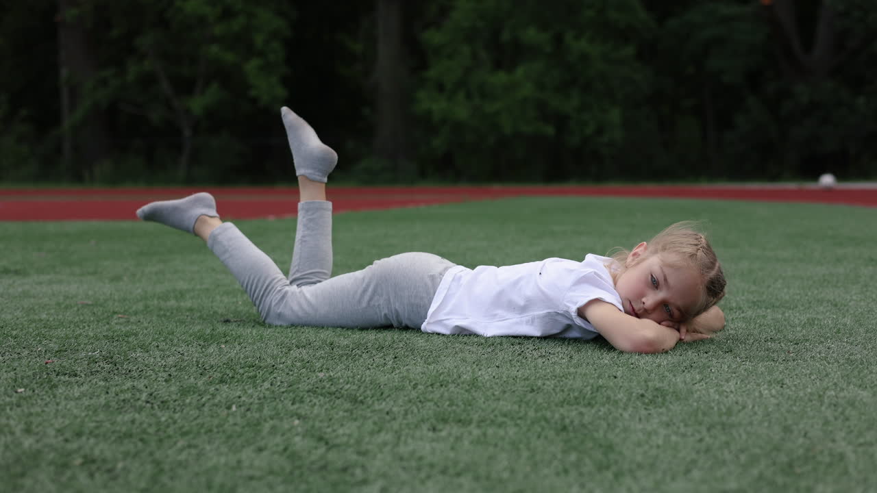 A little girl lies on the green grass and looks at the camera. A stylish girl in a white T-shirt. A girl with braided hair