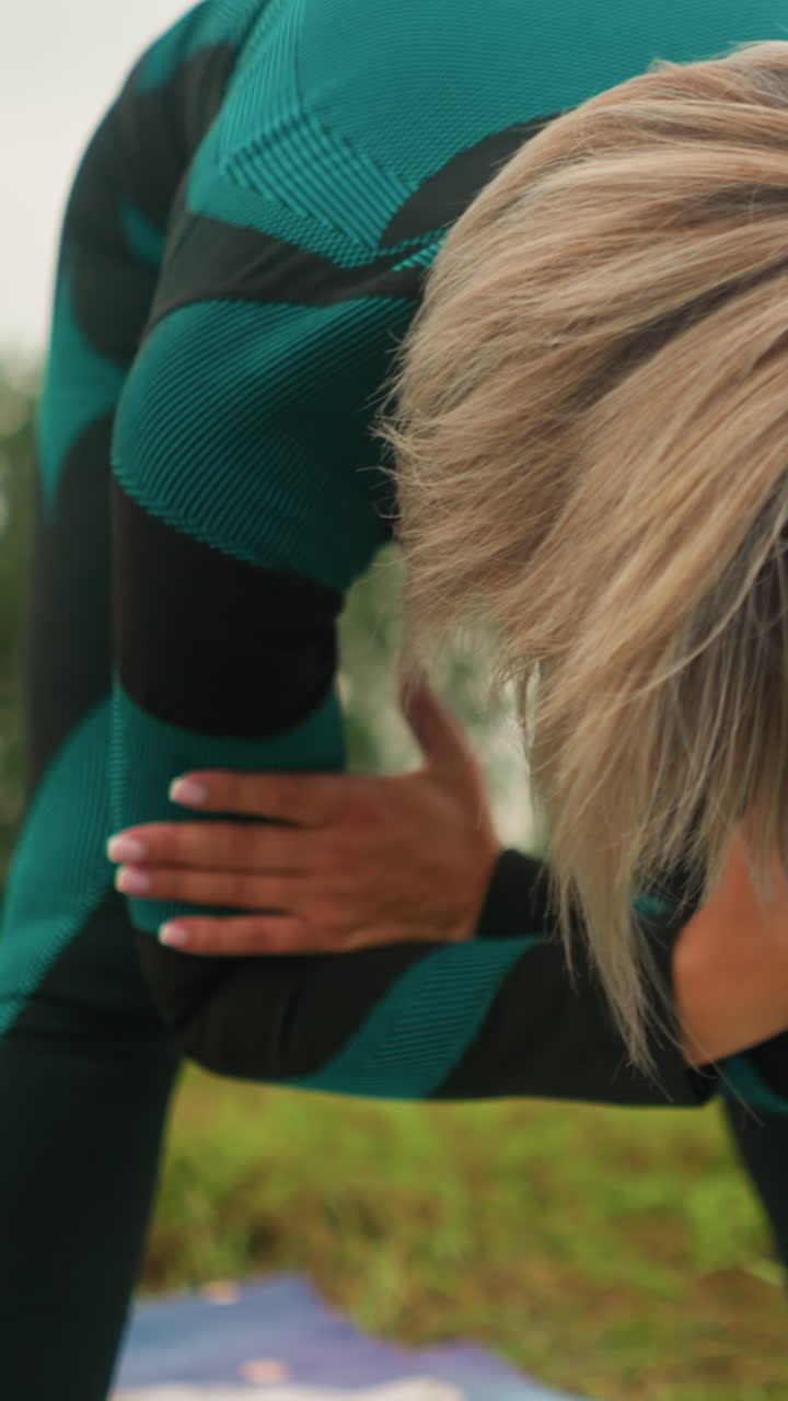 женщина со сложенными руками поворачивается влево и вправо во время практики йоги на огромном травяном поле, ее волосы покрывают ее лицо и размытый коврик для йоги под ней, на заднем плане травяное поле