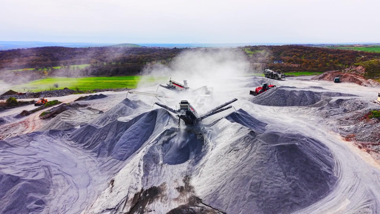 Perspektif drone yang luas dari mesin penyortir yang menghasilkan awan debu di tengah tumpukan kerikil yang luas dekat ladang hijau. Penghancur batu industri beroperasi di lanskap tambang.