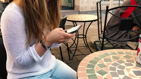 A-young-girl-sits-in-a-chair-at-a-table-outside-a-restaurant-writes-something-on-her-smartphone