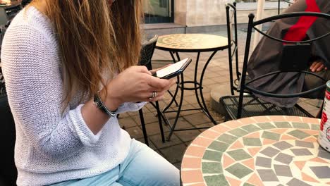 una niña sentada en una silla en una mesa afuera de un restaurante escribe algo en su teléfono inteligente