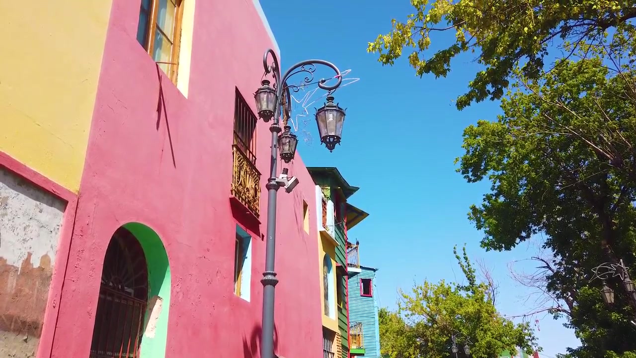 Inclinación de ángulo bajo sobre las coloridas fachadas de Caminito con edificios rosas, amarillos y verde azulado, una farola ornamentada y árboles verdes contra un cielo azul claro en La Boca, Buenos Aires, Argentina.