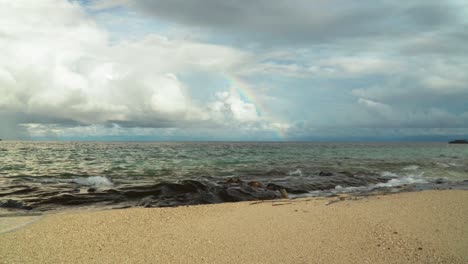 Olas-Rompiendo-En-La-Costa-Rocosa-De-La-Isla-Tropical-Con-Arco-Iris-Y-Nubes-Como-Telón-De-Fondo