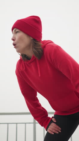 woman in red hoodie and beanie bends down resting, gazing into distance, snowy background with lamp post, frosted trees, benches, iron railing, and mast in foggy winter park atmosphere