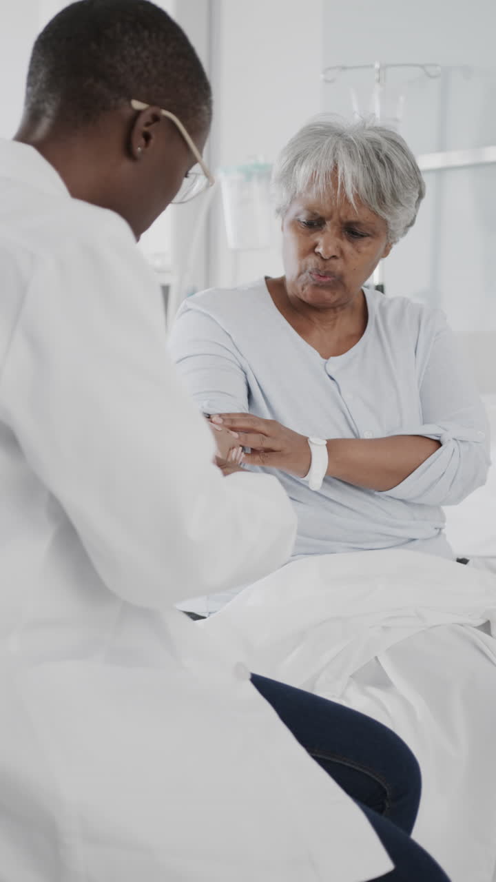 Vertical video of diverse female doctor checking arm of senior female patient, slow motion