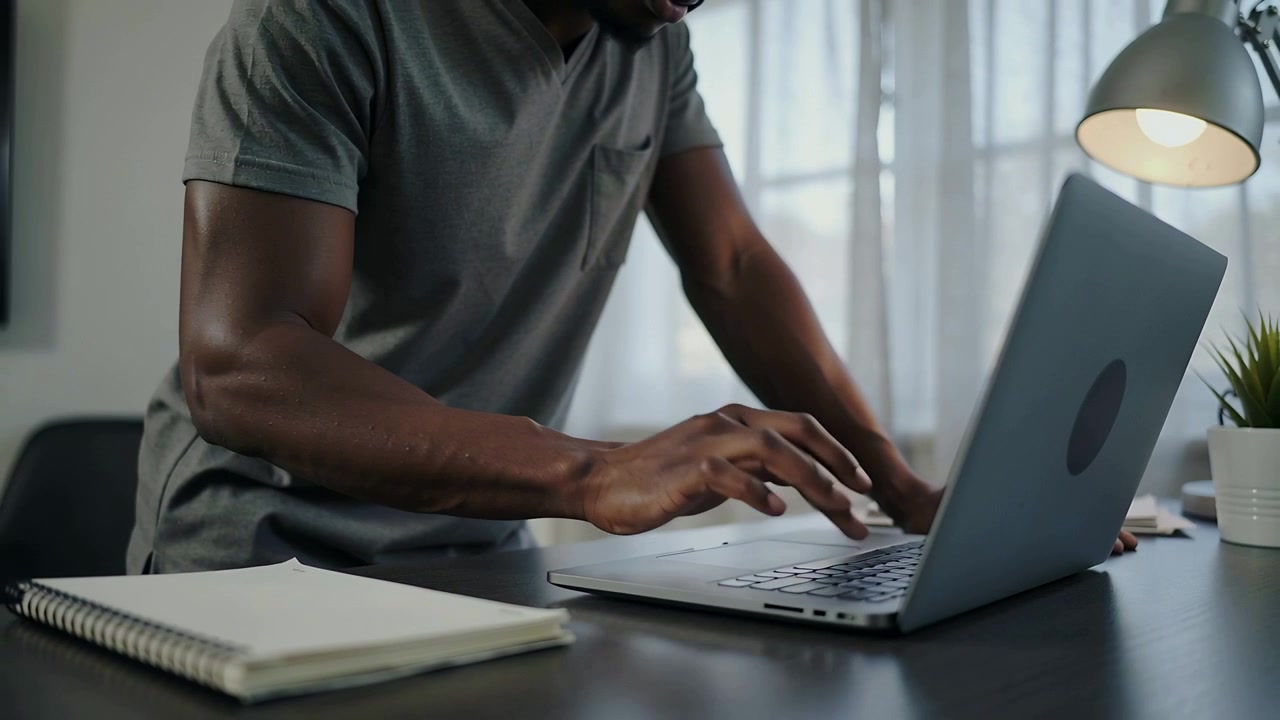 Typing man in gray polo leaning over home desk, prompted by laptop glance, finishing work