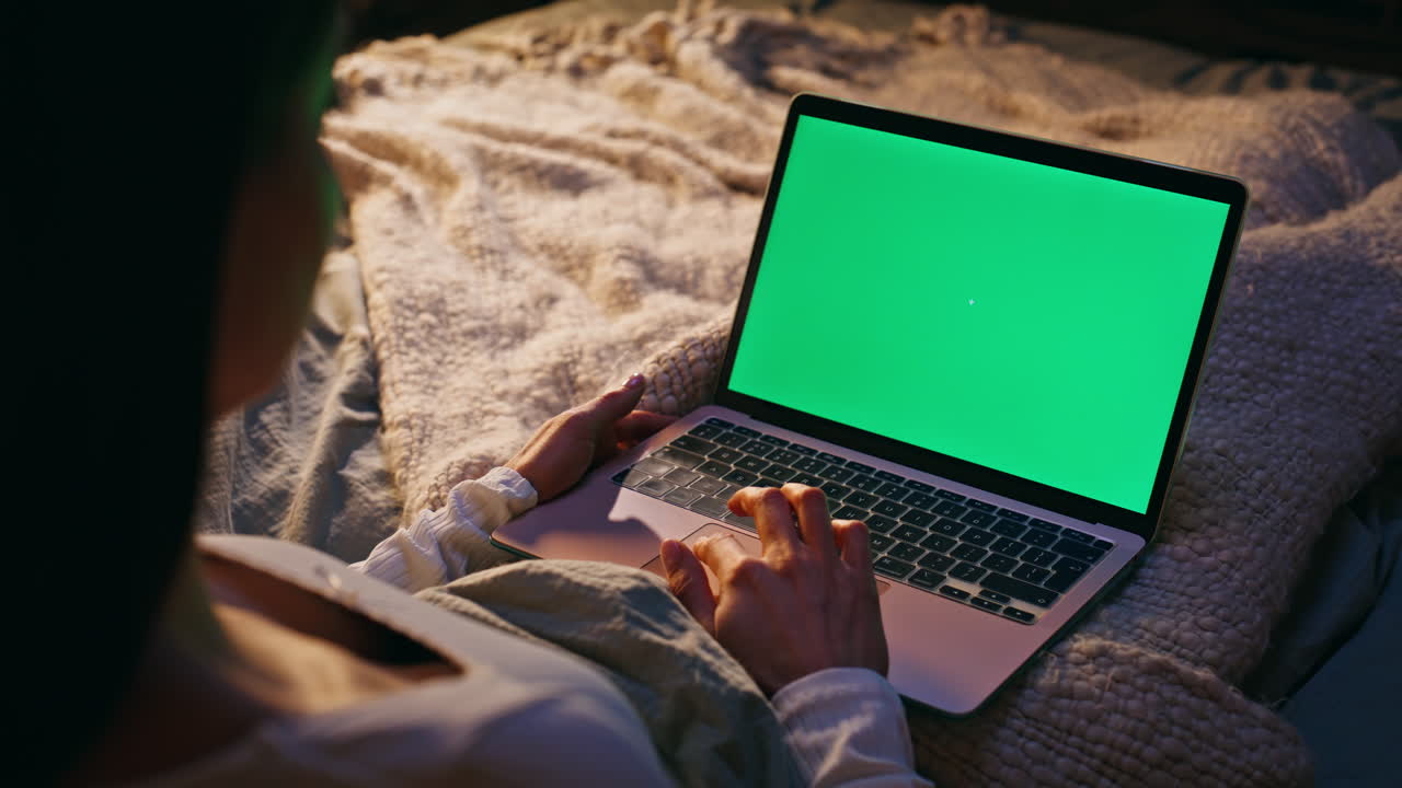 Primo piano di mani di donna che usano il touchpad di un laptop con schermo verde, sdraiata su un letto accogliente a tarda notte.