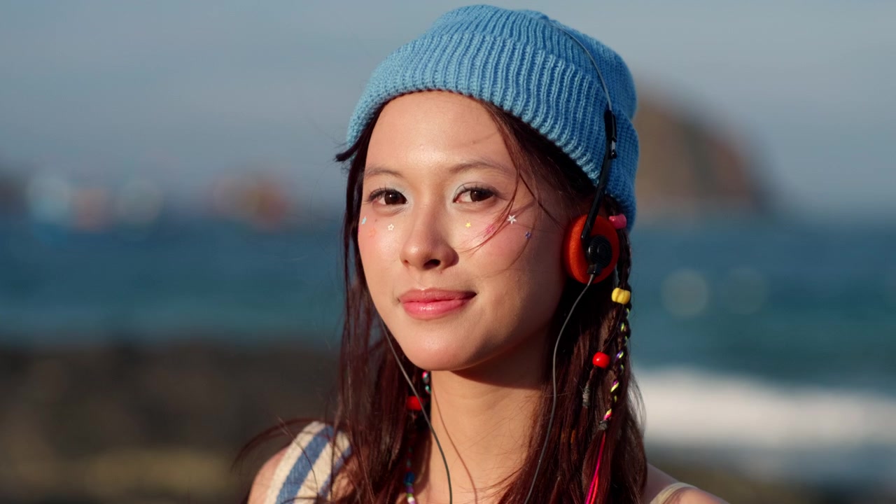 Người phụ nữ châu Á trẻ tuổi đội mũ len xanh nghe nhạc qua tai nghe màu cam bên cạnh đại dương mờ ảo, mỉm cười và nhìn thẳng về phía trước, một chuyến đi ngoài trời để thanh lọc kỹ thuật số