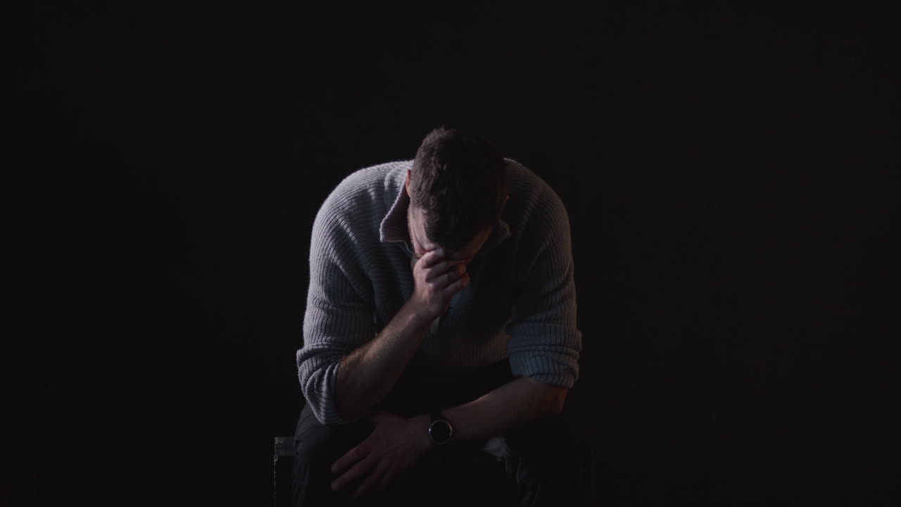Sitting, sad man sobs in dark room, black background