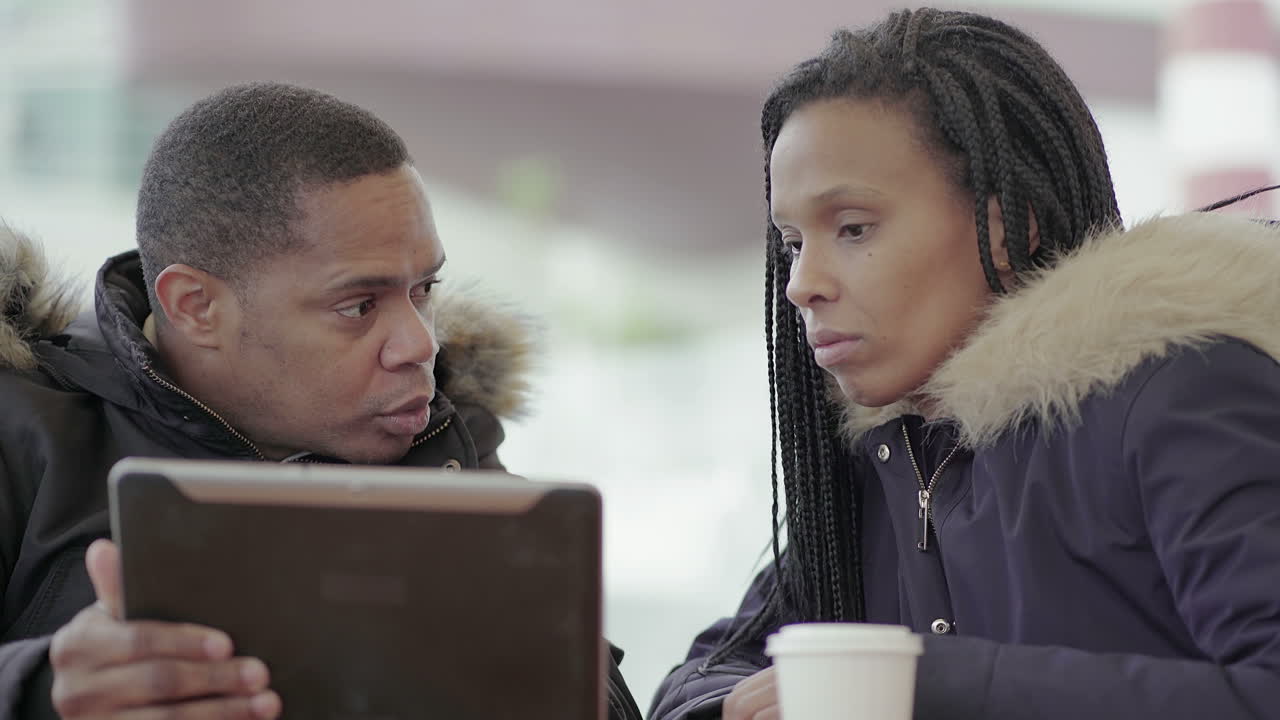 Afro-American middle-aged man in black jacket with fur hood discussing data with Afro-American young girl with braids