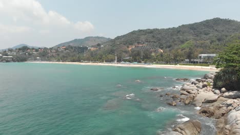 rocky shoreline cliff splitting had karon noi beach from karon beach in phuket, thailand - aerial low angle tracking shot reveal