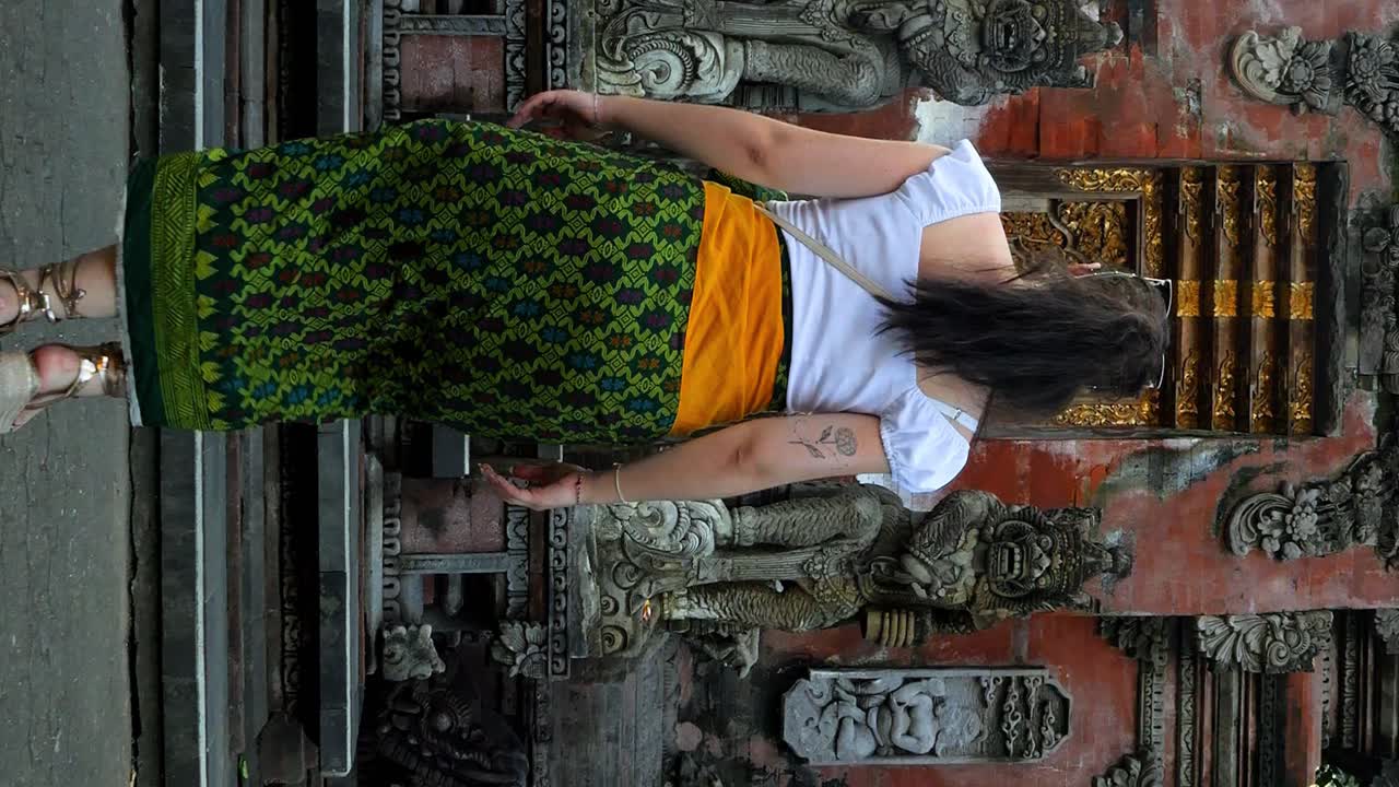 Vertical slow motion dolly shot of a young woman dressed in a green sarong walking towards a temple entrance in bali indonesia