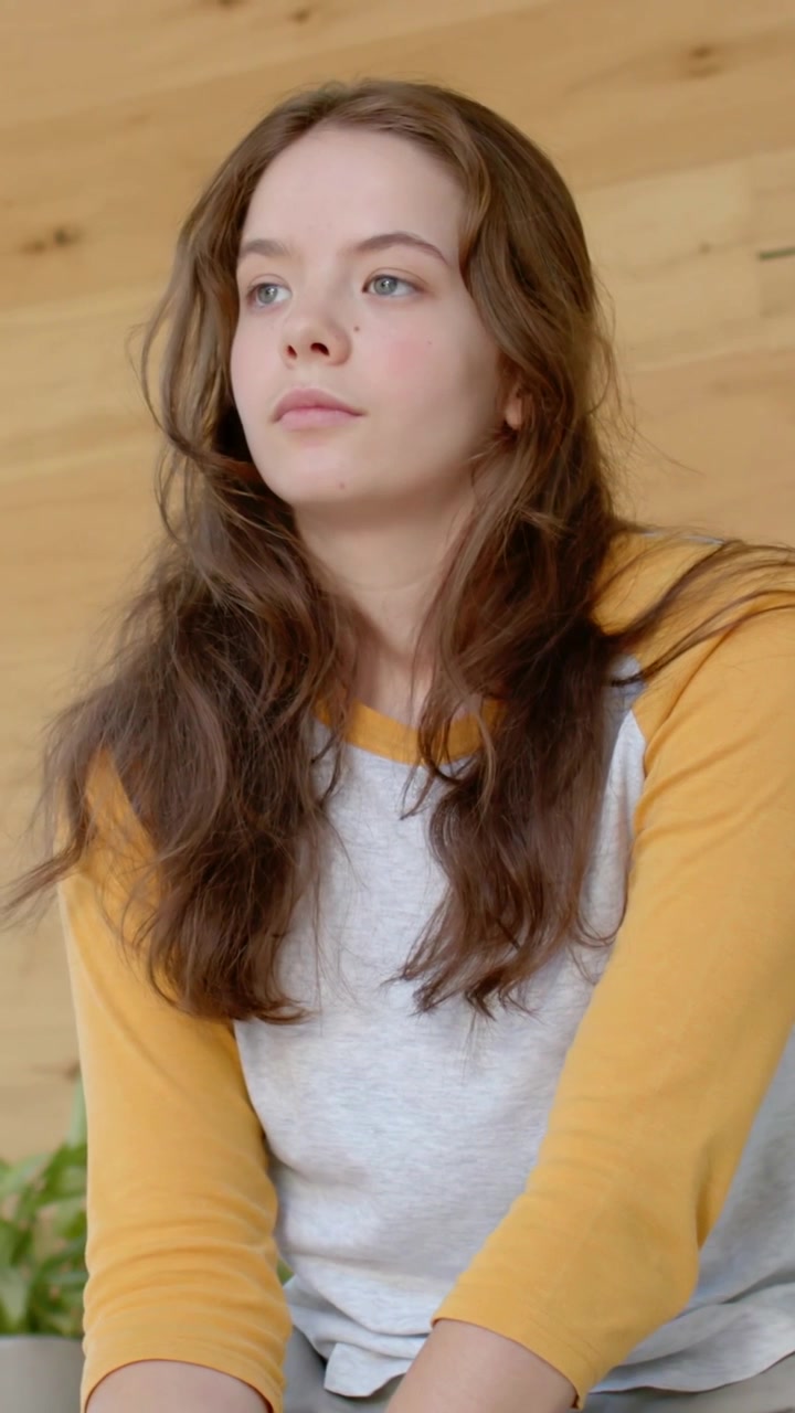 Vertical video: Glancing right, woman yellow raglan turning head, thinking at wood wall, plant left