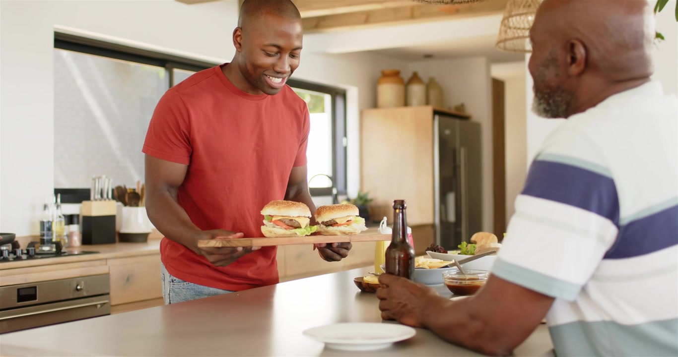 Adult son walking to kitchen island holding wooden tray and offering two burgers for family meal