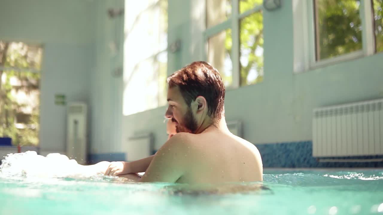 수염을 가진 행복한 젊은 아버지는 닫힌 수영장에서 작은 아들과 함께 시간을 보내고 있습니다. 물 위에서 그를 회전시키고 있습니다. 함께 행복한 시간