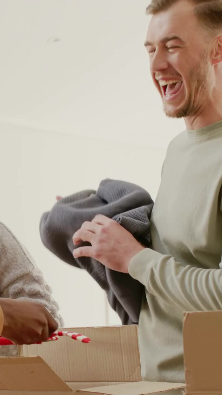 Video verticale: Sollevando il lembo di una scatola, un uomo con una maglia color salvia rimuove un capo in maglia grigio e un peluche rosso, sistemando le cose a casa.