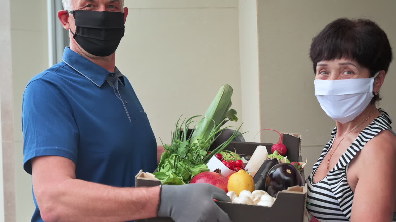 Smiling woman and delivery man wearing masks holding a box of vegetables