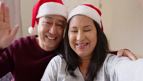 smiling senior asian couple wearing santa hats celebrating christmas at home together