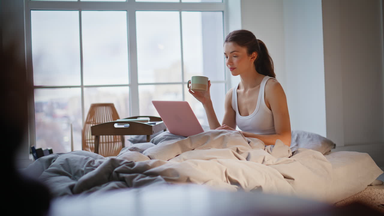 Empresaria mirando el portátil en el dormitorio, bebiendo café y relajándose en una acogedora ropa de cama blanca.
