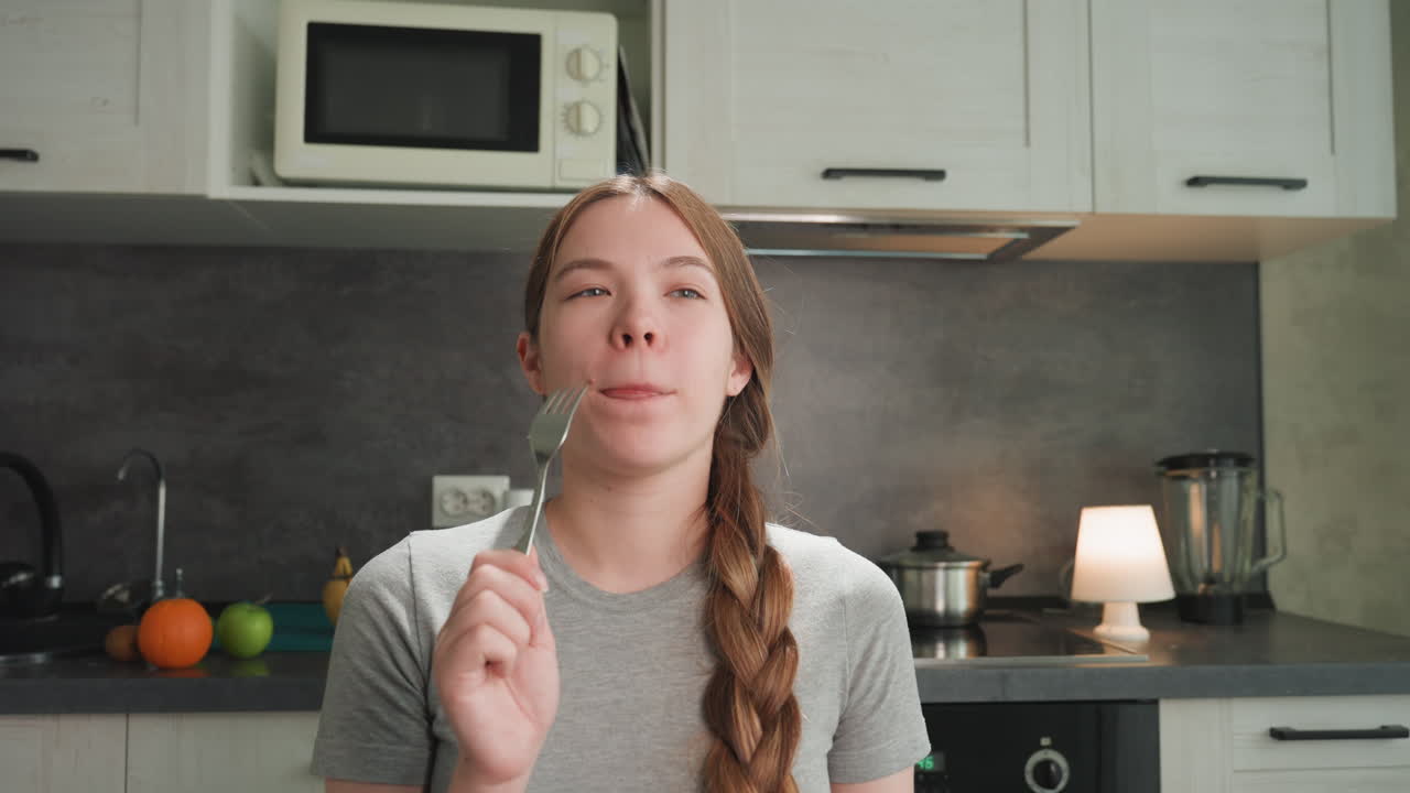 Pregnant white woman tasting fork in kitchen, braided hair, gray tshirt, microwave and blender on counter, bowl of fruit and orange visible, lamp glow, woman sampling food with smile, food blogger