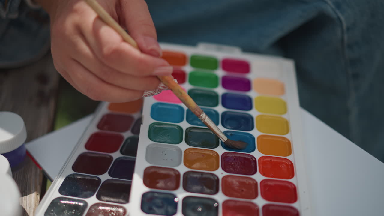 Primo piano di una mano che seleziona pigmenti acquerello vivaci su una tavolozza, con le dita che intingono il pennello in pozzetti di colore intenso. Vivace miscelazione di colori durante una sessione creativa all'aperto. Un'atmosfera gioiosa e sperimentale, con tratti precisi e giocosi.