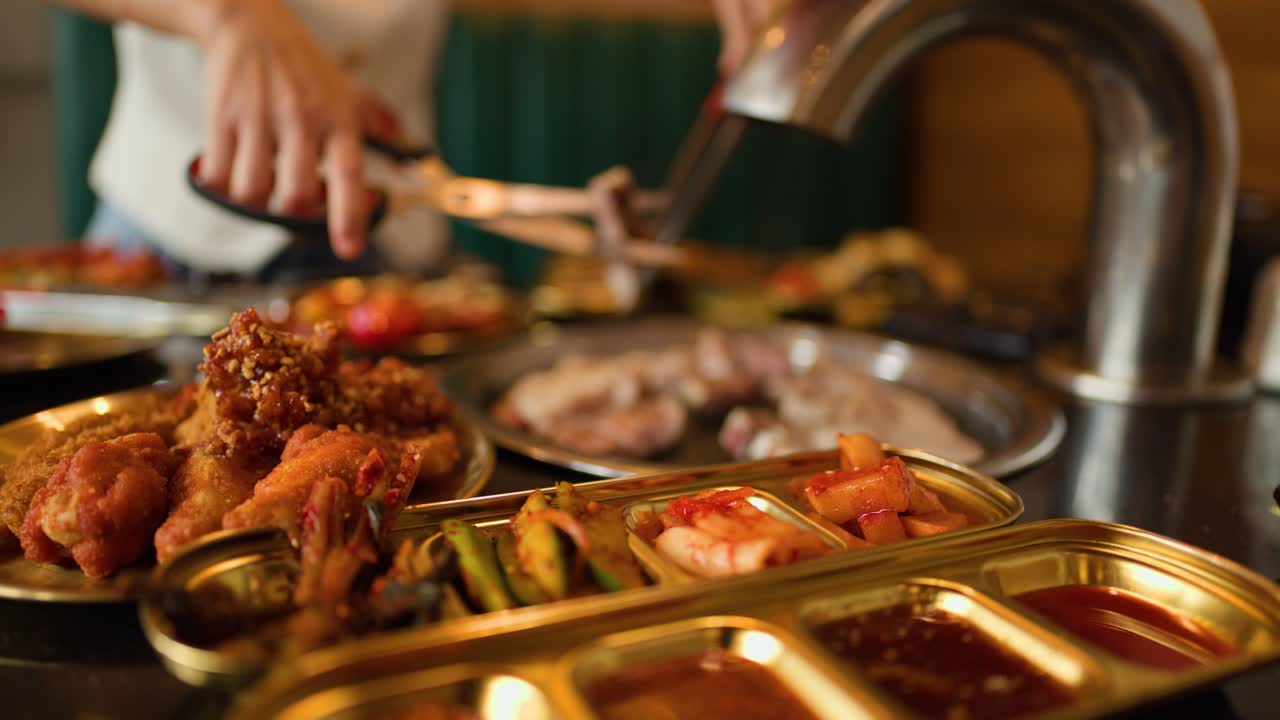 Person cuts grilled meat with scissors at Korean BBQ, golden tray of side dishes visible