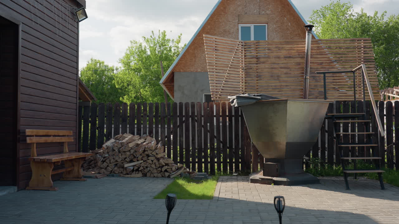 Peaceful view of compound with firewood stacked beside wooden bench near brown fence, large metal tub with chimney and stairs, green trees swaying gently in breeze under bright afternoon light