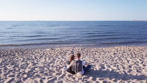 Toma-Aérea-De-Una-Pareja-Joven-Sentada-En-La-Playa-De-Arena,-Abrazándose