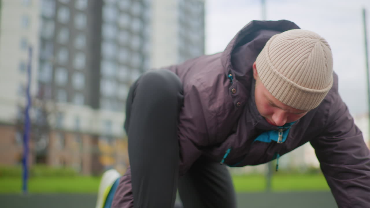 white man tying shoe on urban court, beanie and puffer jacket, basketball in foreground, lowangle closeup, focused expression, apartment towers in background, chilly overcast light, preparing